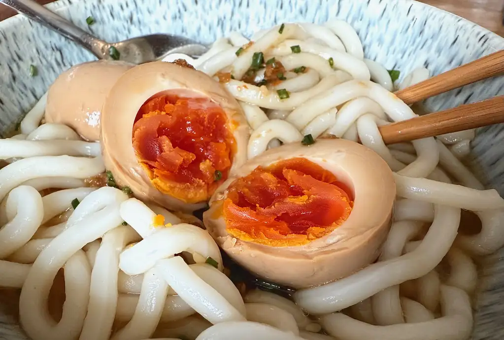 A close-up of a bowl of udon noodles in broth, garnished with finely chopped green onions. Two marinated soft-boiled eggs (mayak gyeran) are halved, revealing their jammy, deep orange yolks. A pair of wooden chopsticks rests on the noodles, and a spoon is partially visible in the background.